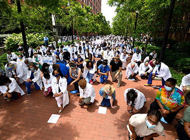 Hundreds of Johns Hopkins clinicians in white lab coats kneel on the campus of the Johns Hopkins hospital.